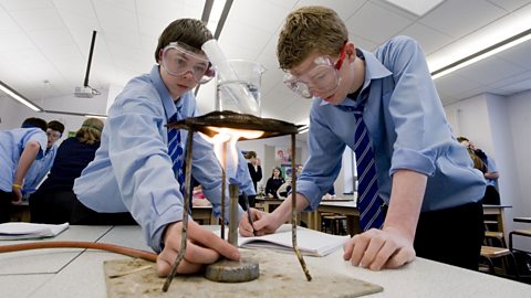 Two school boys conducting experiment using a bunsen burner; both are wearing goggles.