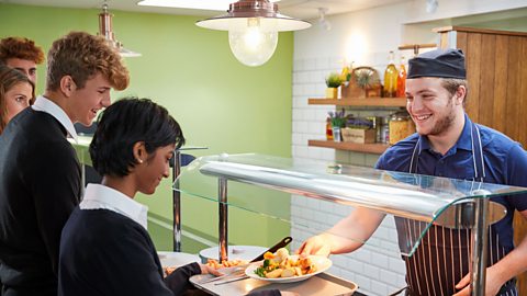 Students line up for food which is served by a chef who is smiling, and hands over a plate of food to the first in the queue.