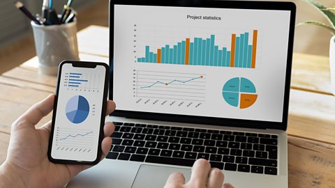 A computer screen with a bar chart and a pie chart on it, as a person holds a mobile in their left hand that also has a pie chart on it.