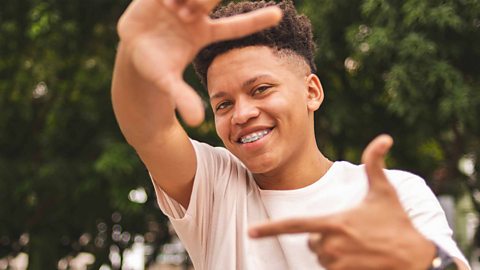 A student looking at camera smiling and holding his hands up as if to frame a photograph. There are trees in the background.