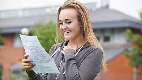 A student standing outside near a building. She is holding a piece of paper which has her exam results on it. She is smiling and holding her chest as if in disbelief.