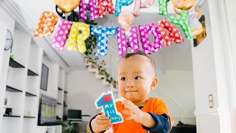 A baby sitting underneath balloons that spell out "happy birthday". The baby is holding a number 1, which says "my 1st birthday".