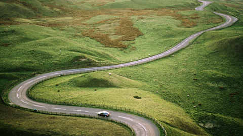 An image from above of a car travelling on a long and winding road surrounded by fields