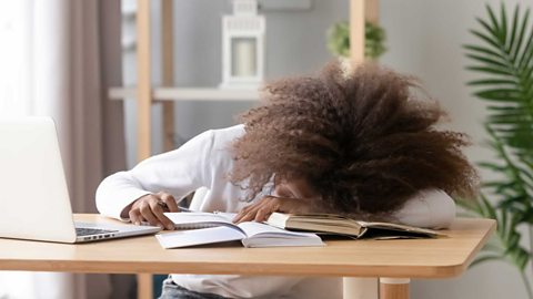 A teenager rests their head on a desk, with books and laptop visible.