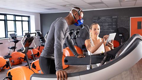 An image of a person undertaking a fitness test; he's wearing a mask whilst standing on a treadmill. A trainer / assessor is by his side monitoring the results.