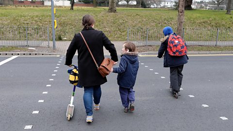 An adult and two children cross a road. The adult and one of the children have scooters.