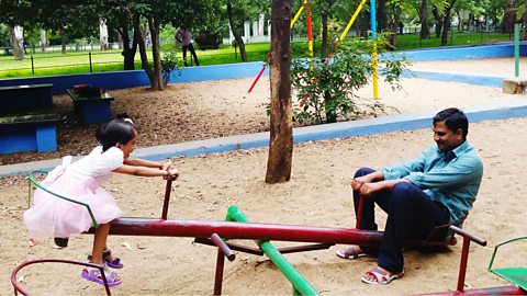 A father and daughter sitting on opposite sides of a seesaw, the daughter lifted into the air and the father near the ground.