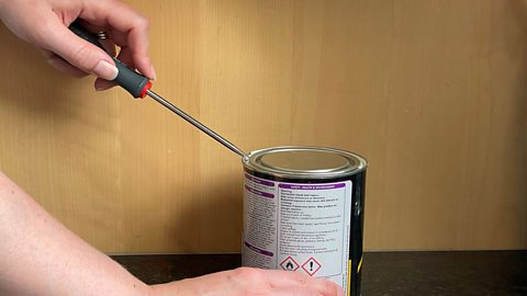 A person using a screwdriver to open a can of paint