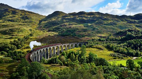 The curve of the Glenfinnan Viaduct with a Jacobite steam train passing over it, with green hills in the background