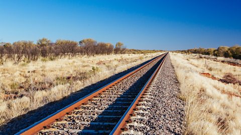 The track of The Ghan railway stretches out to the horizon, with dry shrubs either side and a bright blue sky above