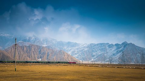 A train on the Qinghai-Tibet railway passes in front of a snow-capped mountain range, underneath a cloudy sky