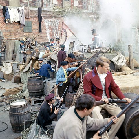 Photograph from the set of Les Miserables, with men holding guns crouching behind the barrels and wood that make up the barricade