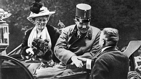 Franz Ferdinand and his wife, Sophie, in their carriage. Sophie is holding a bouquet of flowers, and Franz Ferdinand is shaking hands with a man. 