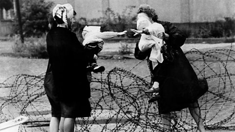 Two women standing in front of barbed wire, holding young children, who are reaching out to touch each other. 