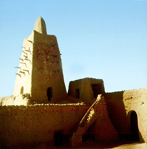 This is the Great Mosque in Timbuktu, built by Mansa Musa. It has a stone tower on top, and it is next to another wall with an entrance cut into it.