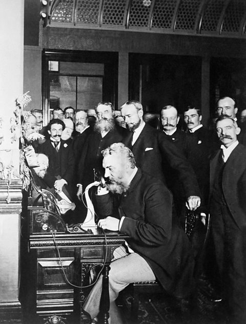 A group of men gathered around Alexander Graham Bell, who is sitting at a desk and talking on a telephone. 