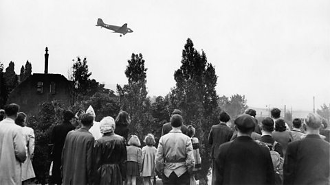 A group of people in West Berlin watch as a US plane flies overhead. 