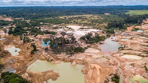 Aerial view of the impact of deforestation on the Amazon rainforest. There are large pools of water and areas of exposed earth. More rainforest can be seen in the background.