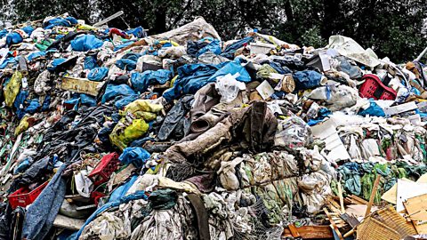 A large pile of mixed waste materials, including plastic bags and fabric, in an outdoor setting.