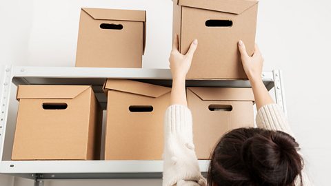 A woman reaching for a cardboard box from the top shelf of a cabinet.