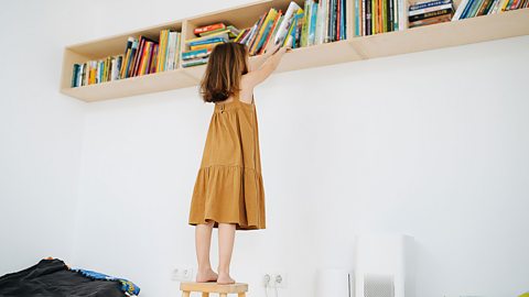 A young girl standing on a stool and reaching for a book from a high bookshelf