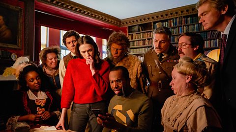 A group shot of the actors from ˿ Ghosts, in costume, gathered around a desk in the library, looking at a phone.