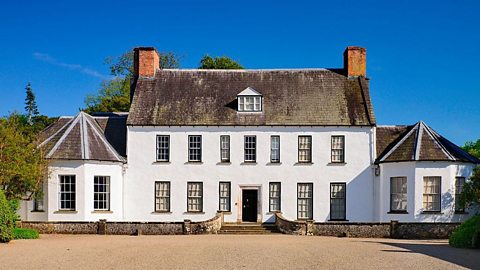 A photograph of the front of Springhill House. The building is painted white and has symmetrical windows and doors. There are rounded rooms on either side of the property with pointed roofs.