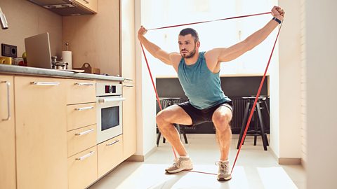 An athletic man crouched exercising with a resistance band in a kitchen.