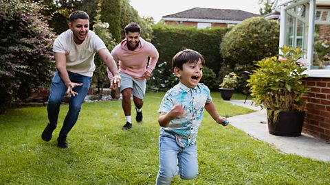 Two dads are chasing their young son around their garden.