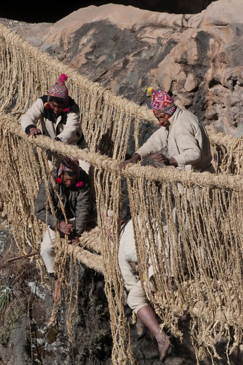 Four men wearing hats and warm clothing stand on or straddle a rope bridge across a canyon, weaving thinner strands of rope into the bridge.