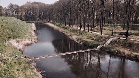 A trench-like bridge cuts through the water of a moat, with trees on the right hand side bank and a steep grassy incline on the left hand side bank,