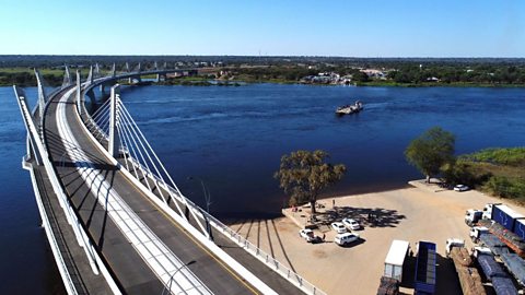 An aerial photograph of the curved Kazungula road bridge, stretching over the Zambezi River. The bridge is empty of vehicles. A boat is on the river.