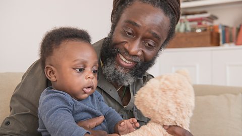 A man with his baby grandson are looking at a teddy bear