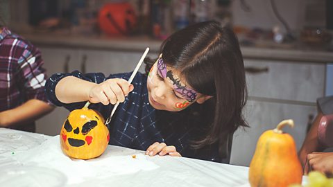 A young girl with face paint and in a witchy Halloween costume is decorating a small pumpkin.