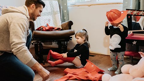 A dad helps his 2 young children to get dressed up as firefighters. Hid little girl sits on the floor and puts on orange tights. The smaller boy is standing up and wearing a firefighter hat.