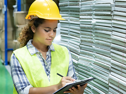 Female in a warehouse wearing a hard hat and hi-viz writing on a clipboard