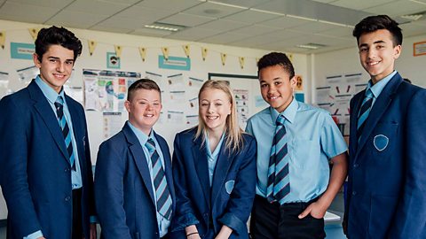 Five students standing in a classroom. The students are looking at camera and smiling. They are wearing blue blazers and shirts.