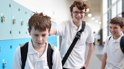 Two students in white polo shirts bullying a third student who looks sad. They are wearing backpacks and standing in front of blue school lockers.