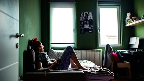 A teenager sits on their bed in a dark bedroom, looking worried and holding their phone.