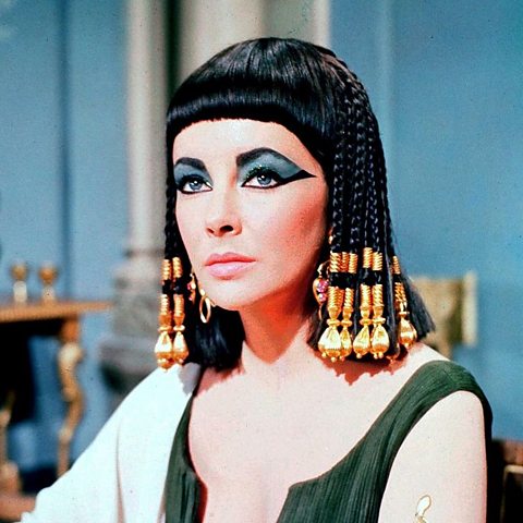 The actress Elizabeth Taylor on set of the film Cleopatra, in 1963. She is wearing a black bobbed wig with gold beads, heavy eye make-up and a green dress.