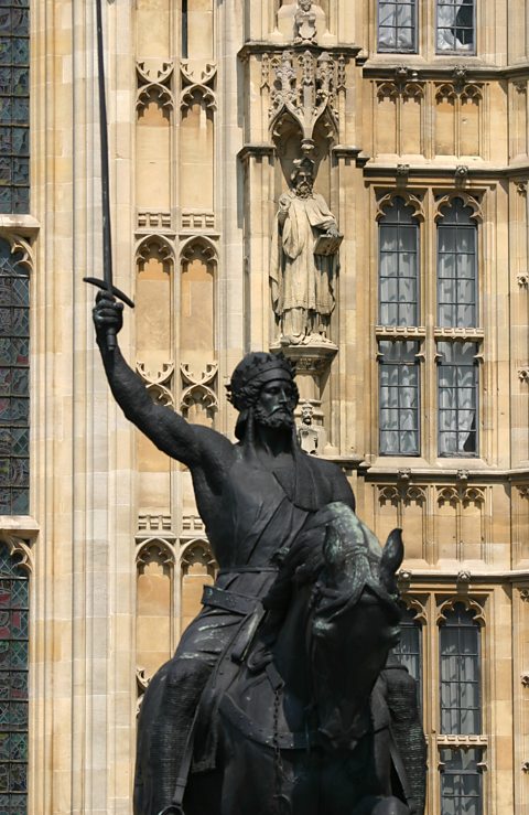 A metal status of King Richard I outside the Houses of Parliament. Richard is sat astride a horse, wearing a crown and chainmail, holding a sword which is raised towards the sky.