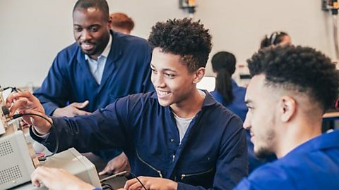 Two students and their teacher, all in blue overalls, using some IT equipment in a classroom.