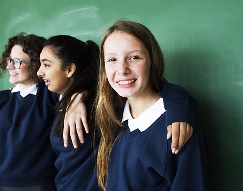 Three teenagers in a school uniform. They are standing in front of a chalk board with their arms around their shoulders.