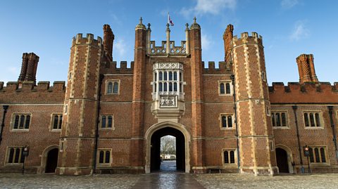 The front of Hampton Court Palace. The building is made of red brick and there are two turrets either side an arched entranceway.