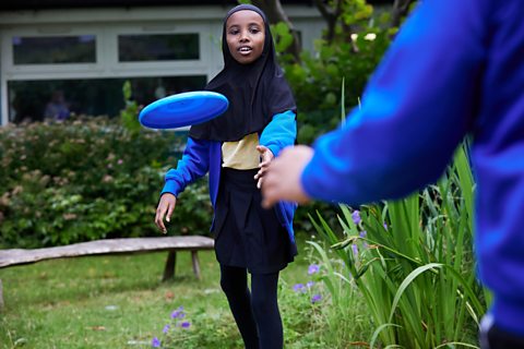 A young girl plays frisbee with her friend, outside in the school garden.