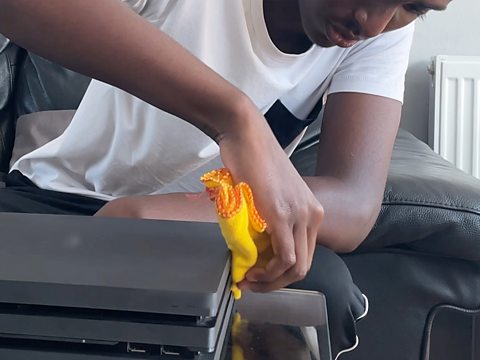 A young man dusts down his games console using a cloth