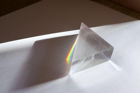 A glass prism on a white surface dispersing sunlight into a rainbow spectrum of colours