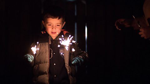 A young boy in a big coat and wearing gloves is holding 2 sparklers in his hands.