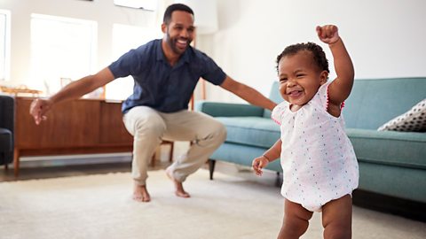 A dad and his young baby daughter are dancing in the living room.