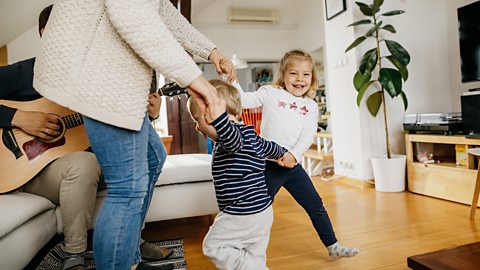 A family of four are dancing in their living room - the dad is playing a guitar.